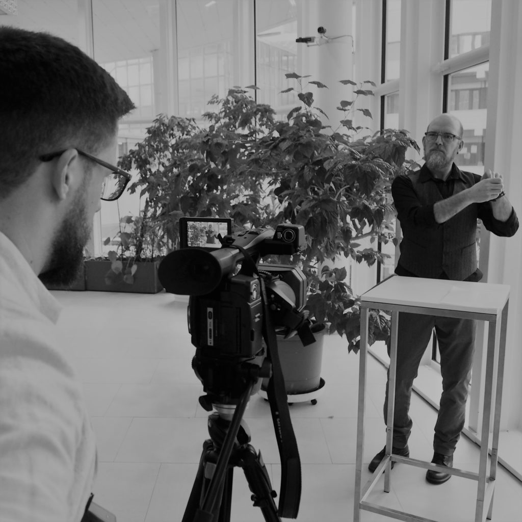 Filming for a career guidance video about Deaf interpreters. One man looks into the screen of a camera on a tripod, filming an iterview with another man who is signing his responses.